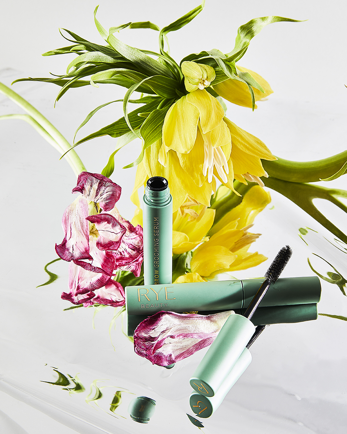 Product shot of RYE Beauty eyebrow grooming serum and mascara, with pink and yellow flowers on a reflective white surface.