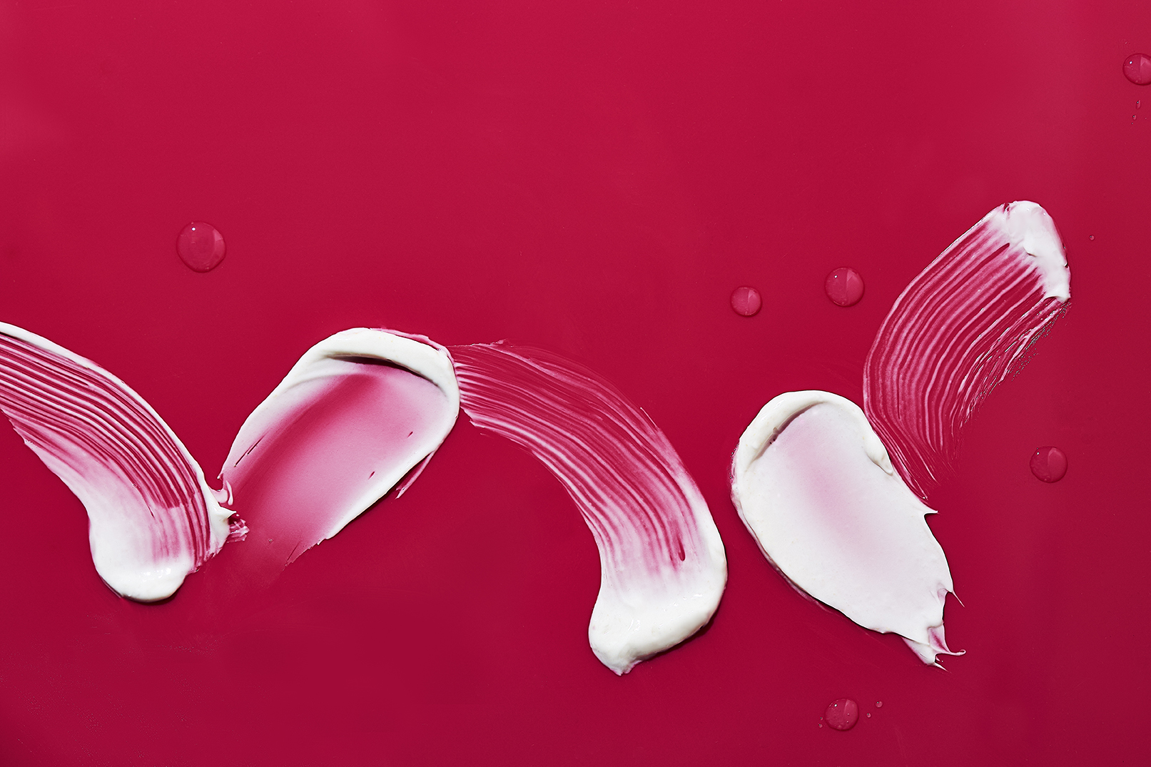 Creamy smears of lotion or skincare product are displayed on a vibrant red background, with tiny droplets scattered around the smears.