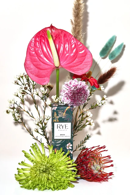 A product shot of Rye Beauty Bloom+ Clarifying Serum featuring pink Anthurium, Chrysanthemum, and assorted flowers on a white background.