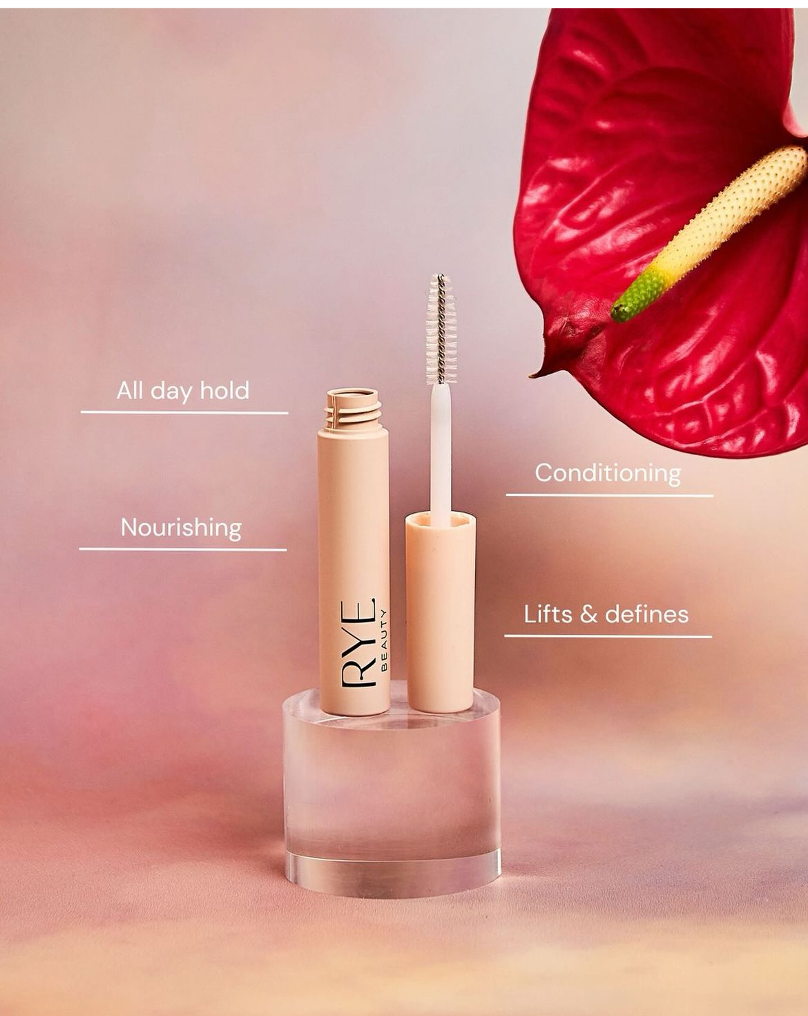 Beige tube of RYE Beauty mascara with applicator, resting on a clear cube with a red anthurium flower in the peach gradient background.