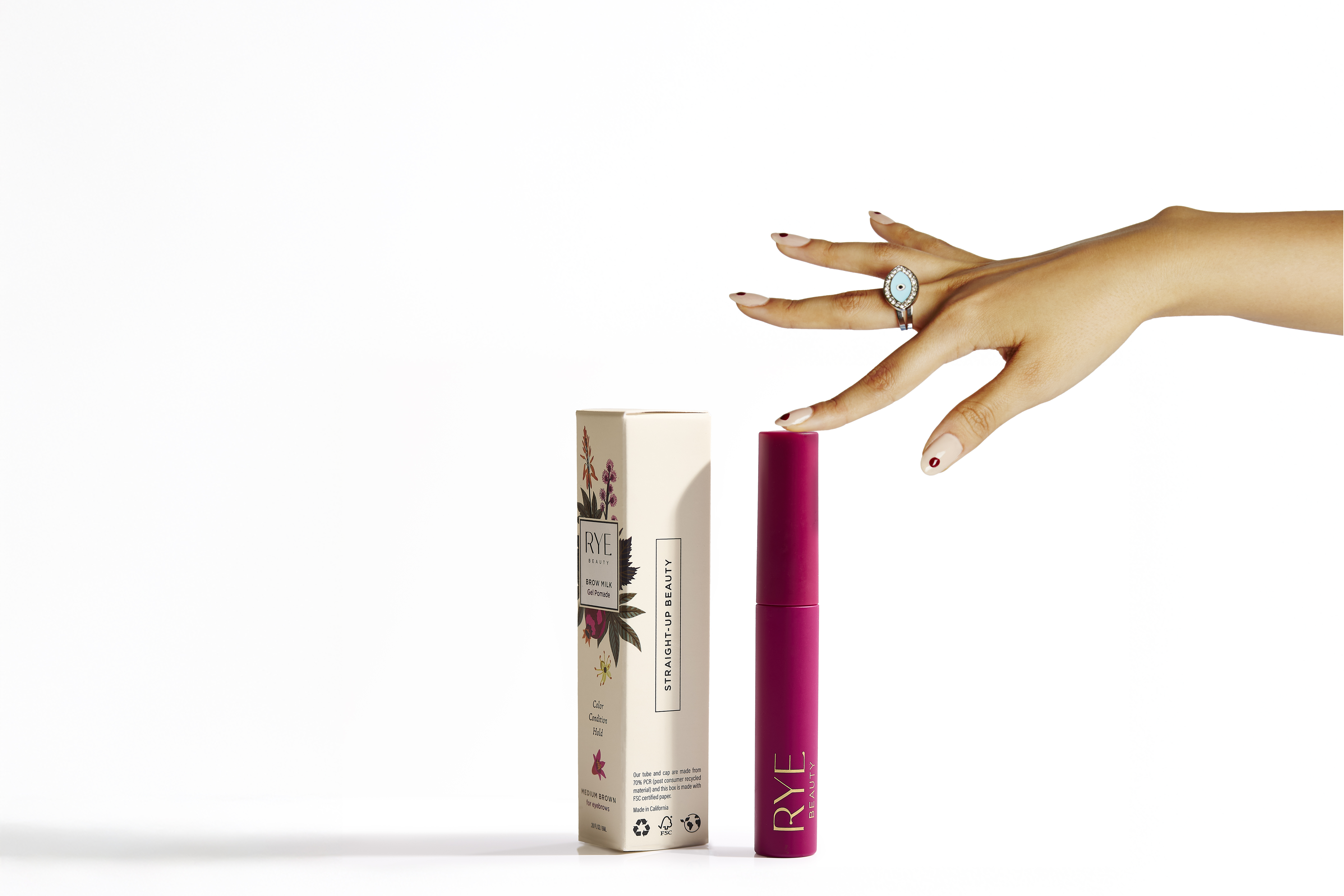 Product shot of Rye Beauty's 'Straight-Up Beauty' mascara with a light-skinned hand wearing a large blue ring on a white background.