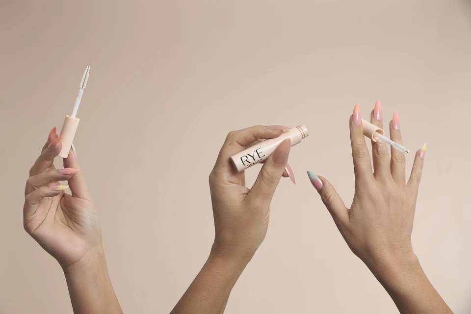 Close-up of three hands holding Rye Beauty eyelash enhancing serum against a light beige backdrop. Focus on product and presentation.