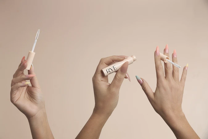 Close-up of three hands holding Rye Beauty eyelash enhancing serum against a light beige backdrop. Focus on product and presentation.