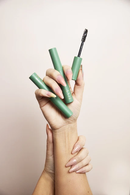 Close-up of hands holding green mascara tubes, one open with applicator. Beauty and makeup product promotion against a pale backdrop.