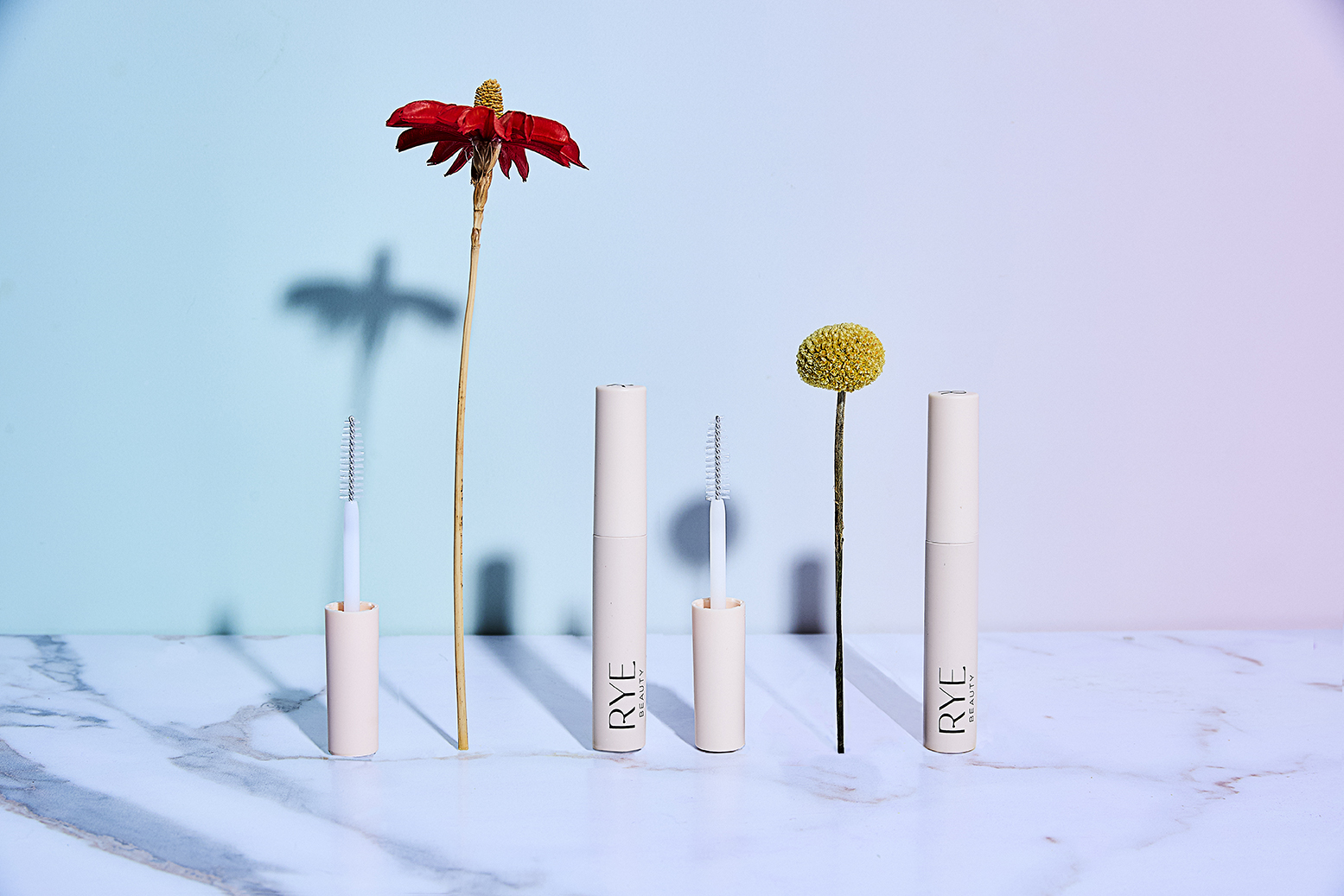Cosmetic product tubes arranged with flowers in a studio setting with a blue and pink gradient backdrop and marble surface.
