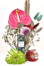 Rye Beauty Brow Conditioning Serum box standing amidst colorful floral blooms including pink anthurium and green spider mum.