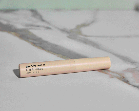 A beige Brow Milk Gel Pomade tube resting horizontally across a white and grey veined marble surface in soft light.