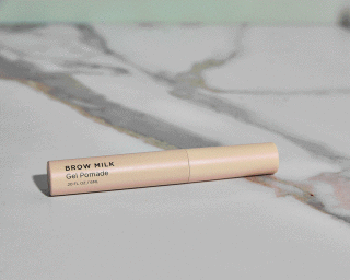 A beige Brow Milk Gel Pomade tube resting horizontally across a white and grey veined marble surface in soft light.