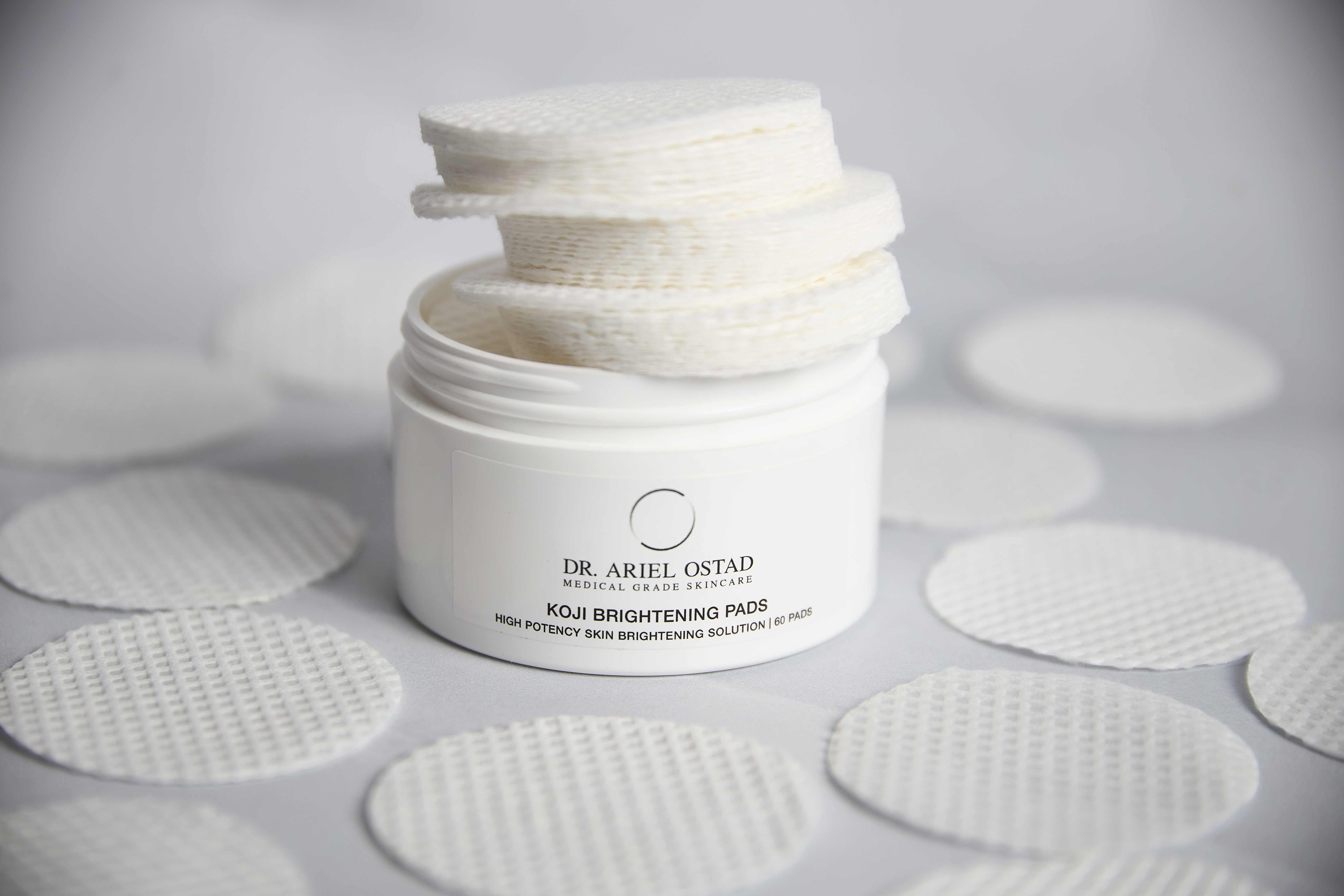 Product shot of a white jar of Dr. Ariel Ostad Koji Brightening Pads surrounded by loose pads on a white surface.