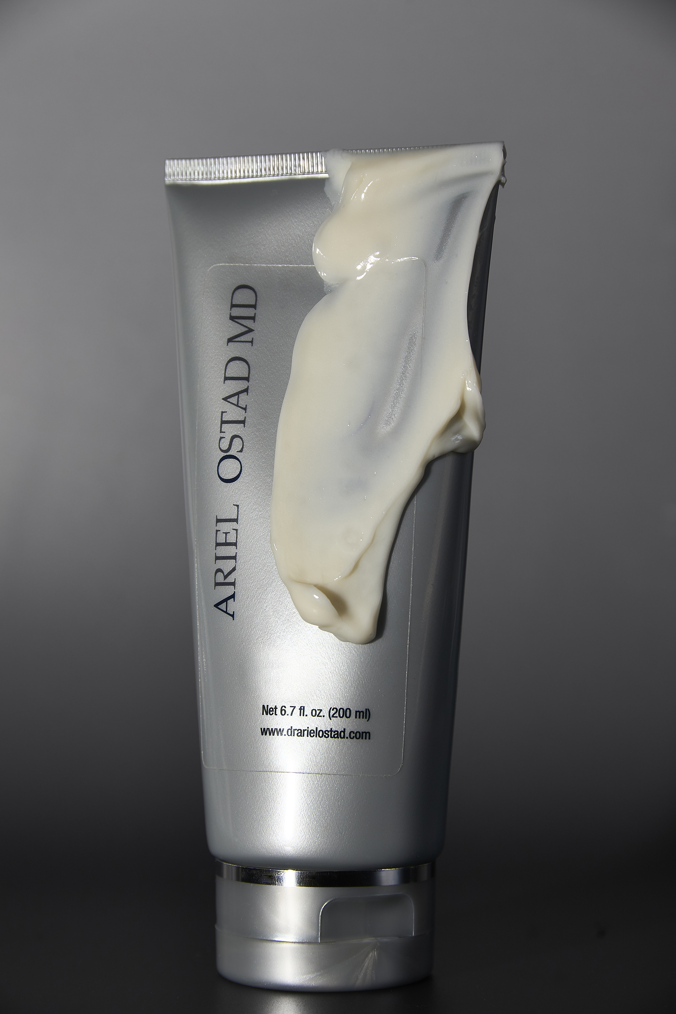 Close-up of a silver tube of Ariel Ostad MD skincare cream with a blob of the product on the side against a grey background.
