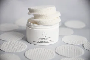 An open white jar of Dr. Ariel Ostad Koji Brightening Pads stacked high with textured cotton pads on a light grey studio surface.