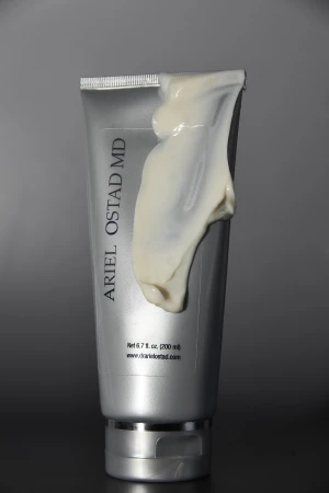 A silver Ariel Ostad MD skincare tube coated with a thick smear of cream lotion against a gradient grey background.