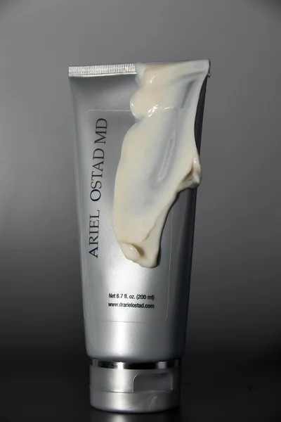 A silver Ariel Ostad MD skincare tube coated with a thick smear of cream lotion against a gradient grey background.