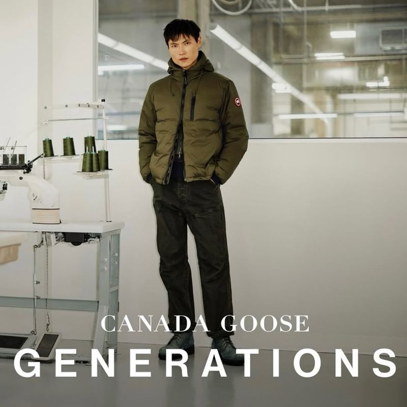 A full-length shot of a stylish adult man wearing a hooded puffer jacket, jeans, and sneakers, standing indoors with natural light.