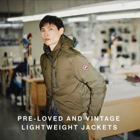 An Asian man wearing an olive green hooded puffer jacket inside a bright clothing manufacturing space featuring sewing and cutting tables.