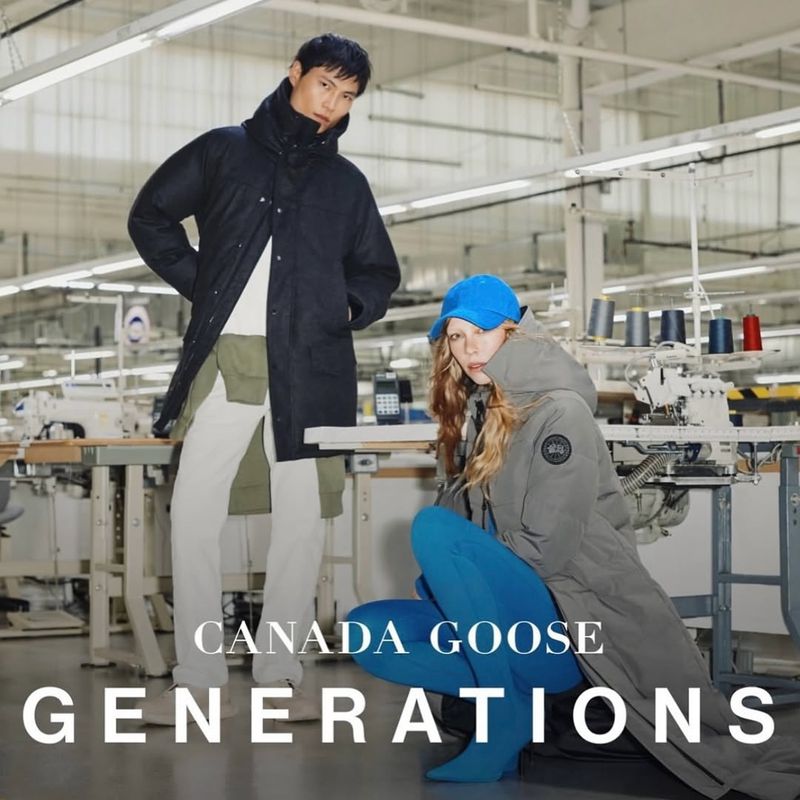 Two adults pose indoors wearing Canada Goose outerwear, one standing in a long dark coat, the other crouched in a light-colored parka.