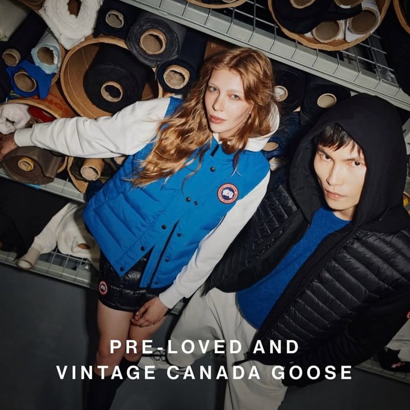 Two people pose in a fashion shot showcasing Canada Goose apparel; a blue vest and black jacket, against a background of fabric rolls.