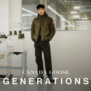 Model standing in an olive green puffer jacket and dark trousers inside a workshop setting with a sewing machine.