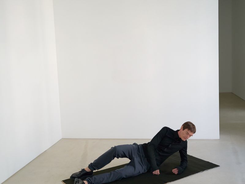 Man in athletic wear lounges on black mat in bright minimalist space with white walls and light grey floor, showcasing casual pose.