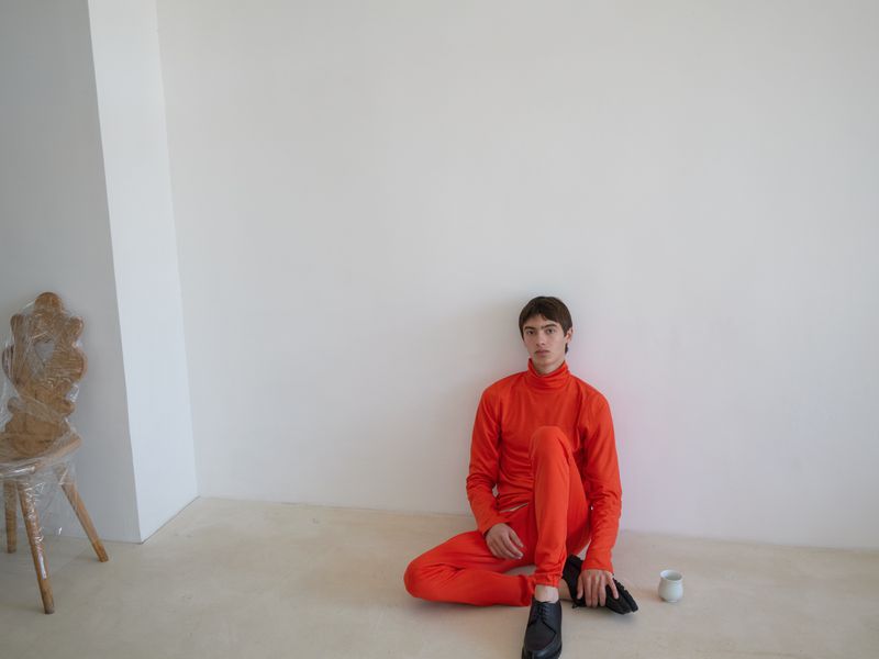 A young adult sits on the floor wearing an orange jumpsuit indoors, against a white wall with a vintage chair and a cup nearby.