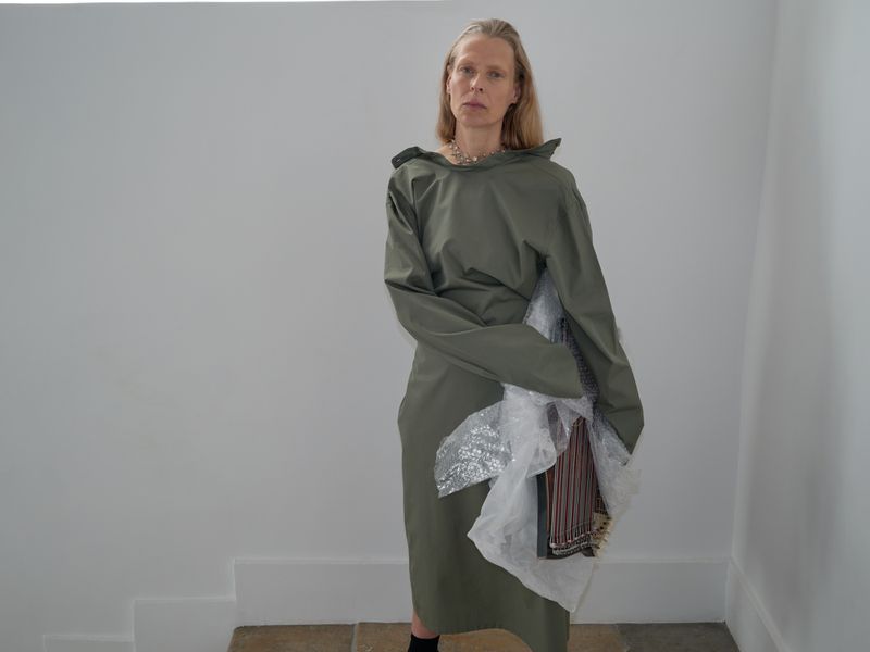 Adult female in an olive green dress stands in a brightly lit interior holding wrapped fabric samples, looking straight at the camera.