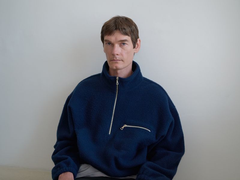 Man in a navy blue fleece jacket with a zipper stands indoors against a neutral wall, captured in a portrait framed at eye-level.