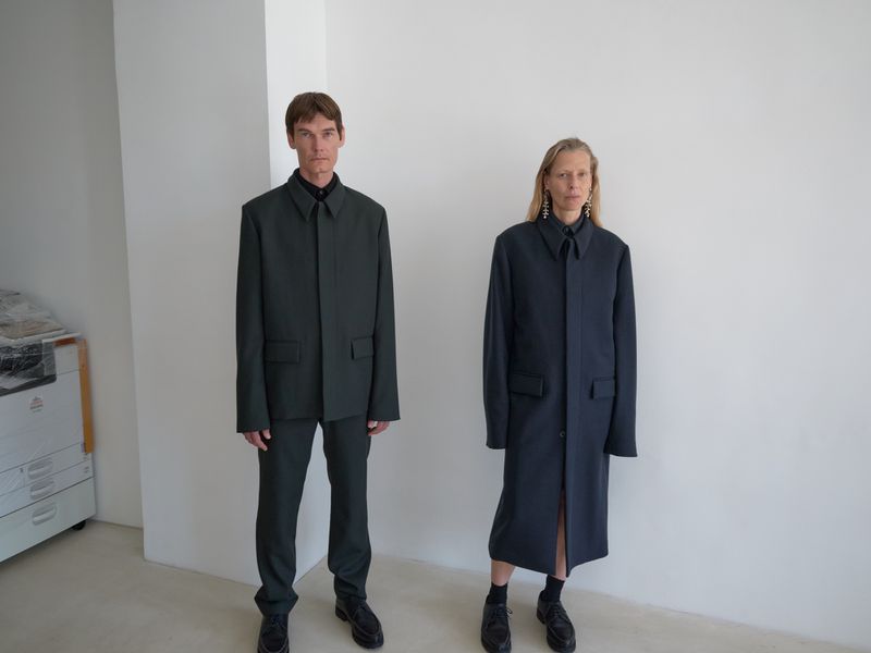 Two fashion models pose in a minimalist studio setting. The adult male wears a green suit, and the adult female wears a blue coat.