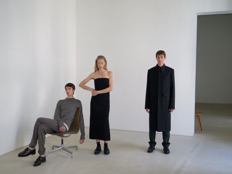 Three fashion models pose in a stark white studio. A woman stands wearing a black dress, and two men wear minimalist clothing. 