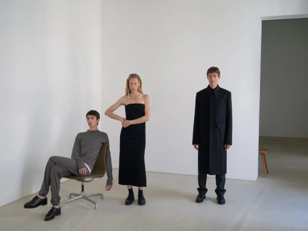Three fashion models pose in a stark white studio. A woman stands wearing a black dress, and two men wear minimalist clothing. 