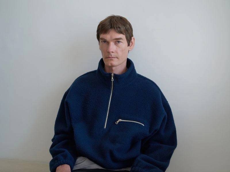Portrait of a male model wearing a dark blue zip-up fleece jacket against a plain light wall.