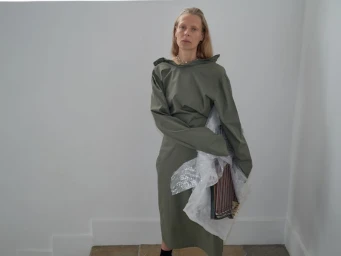 Model in olive green dress holding wrapped item against a plain interior background