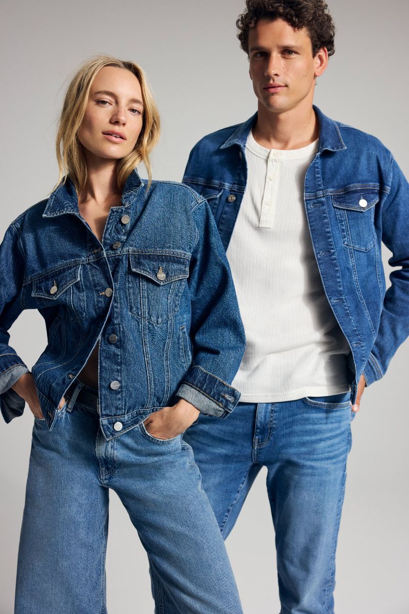 Two fashion models pose in denim jackets and jeans in a studio shot. The woman has blonde hair, and the man has dark curly hair.