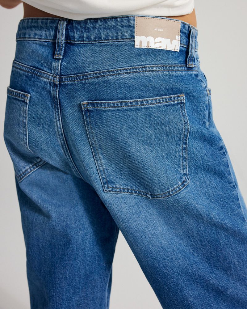 Close-up of a person wearing blue jeans with back pocket detail and a brand tag on the waistband against a neutral background.
