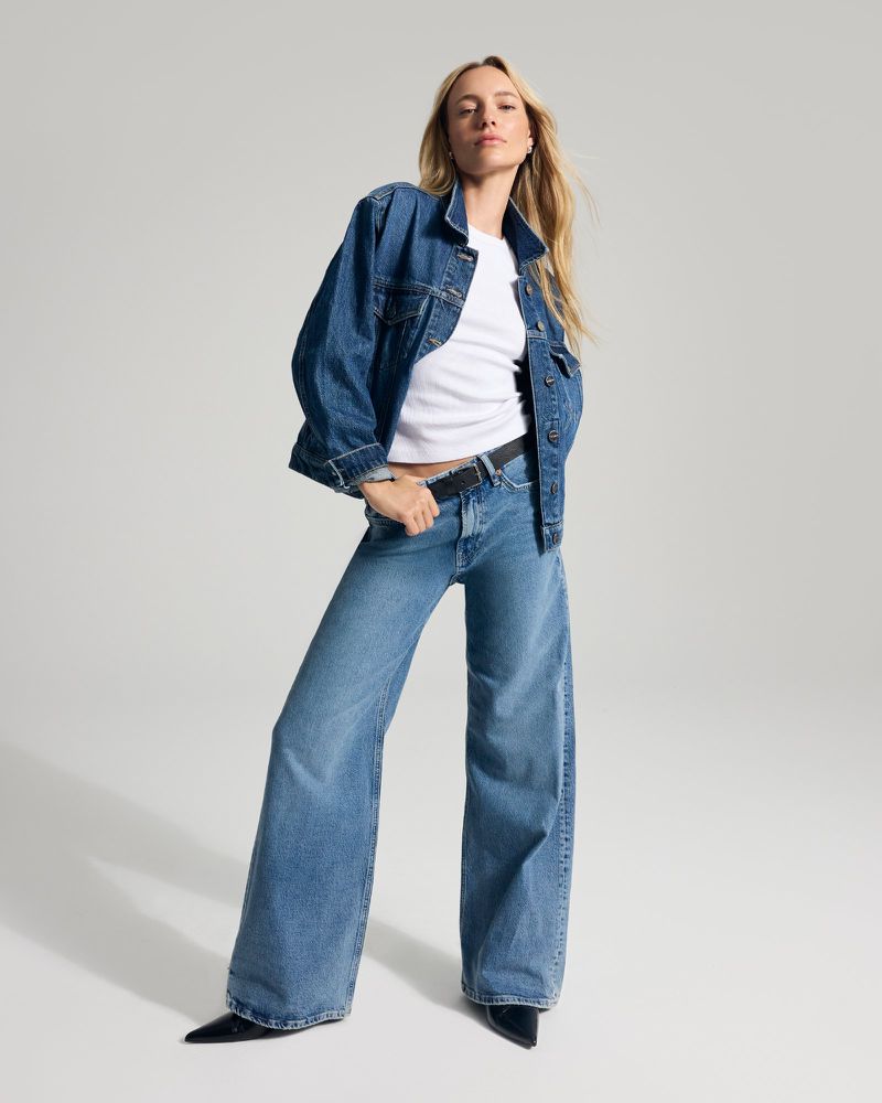 Full-length shot of a blonde woman in denim jacket, white t-shirt, and wide-leg jeans posing in a photo studio with a neutral backdrop.