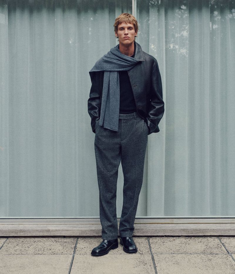 Full-length shot of an adult male fashion model wearing a gray scarf, black jacket, gray pants, and black shoes against a light background.