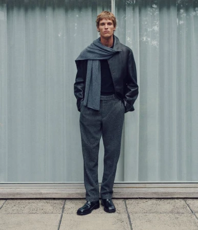 Full-length shot of an adult male fashion model wearing a gray scarf, black jacket, gray pants, and black shoes against a light background.