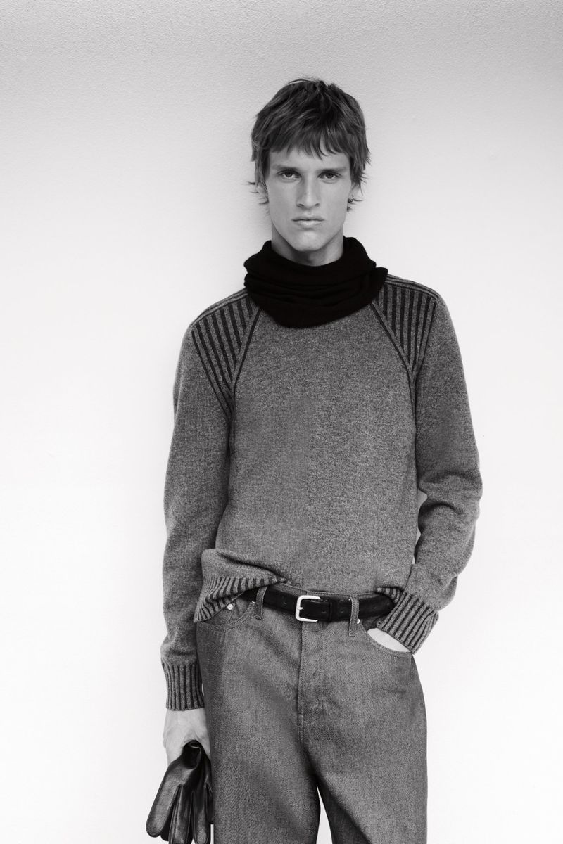 Black and white portrait of a blond male model wearing a gray sweater and black scarf, posing against a plain wall, holding gloves.