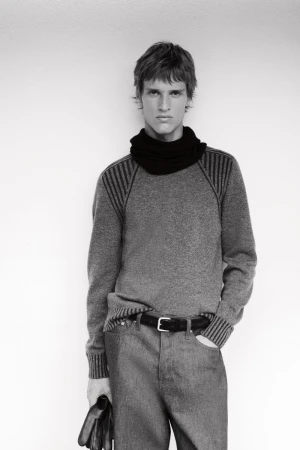Black and white portrait of a blond male model wearing a gray sweater and black scarf, posing against a plain wall, holding gloves.