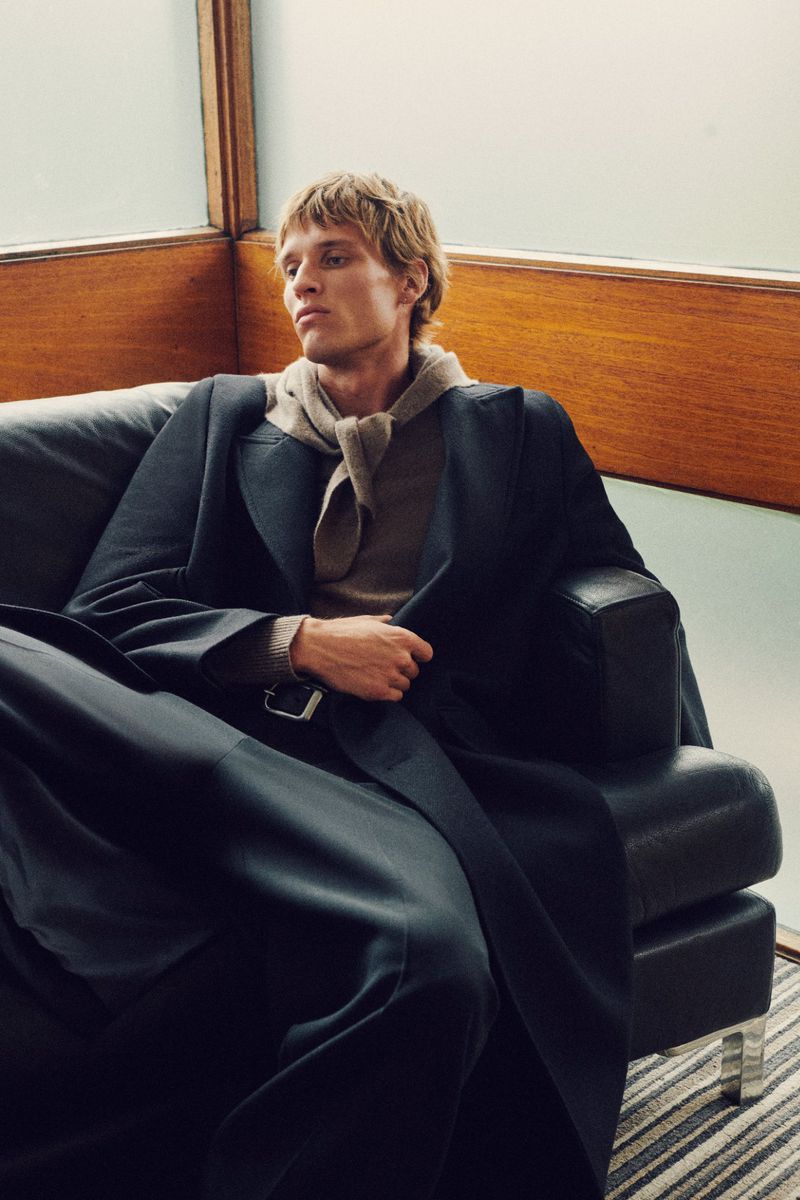 A fair-skinned adult male with blonde hair sits on a black leather couch wearing a black overcoat, brown sweater, and dark pants indoors.
