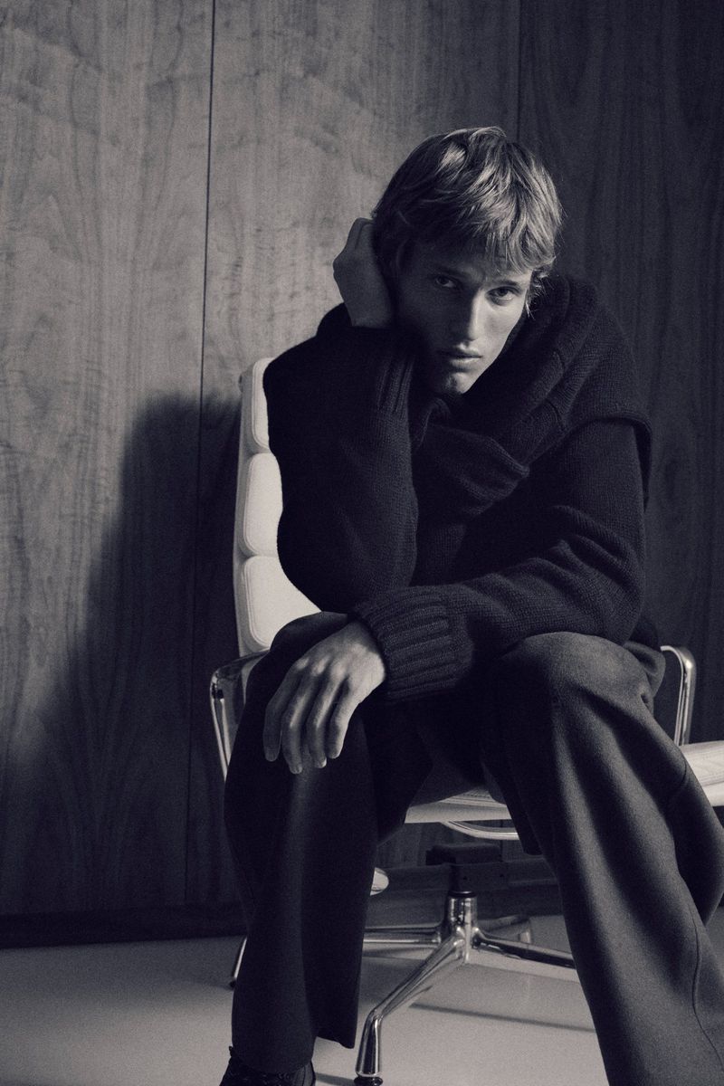 A young adult man poses in a dimly lit studio, seated in a white office chair against a wood paneled wall, wearing a dark sweater.