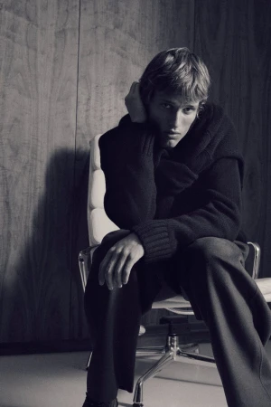 A young adult man poses in a dimly lit studio, seated in a white office chair against a wood paneled wall, wearing a dark sweater.
