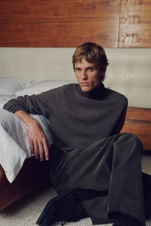 Man in grey turtleneck sweater and pants sits on floor in front of bed, lit by soft, warm light in an indoor environment.