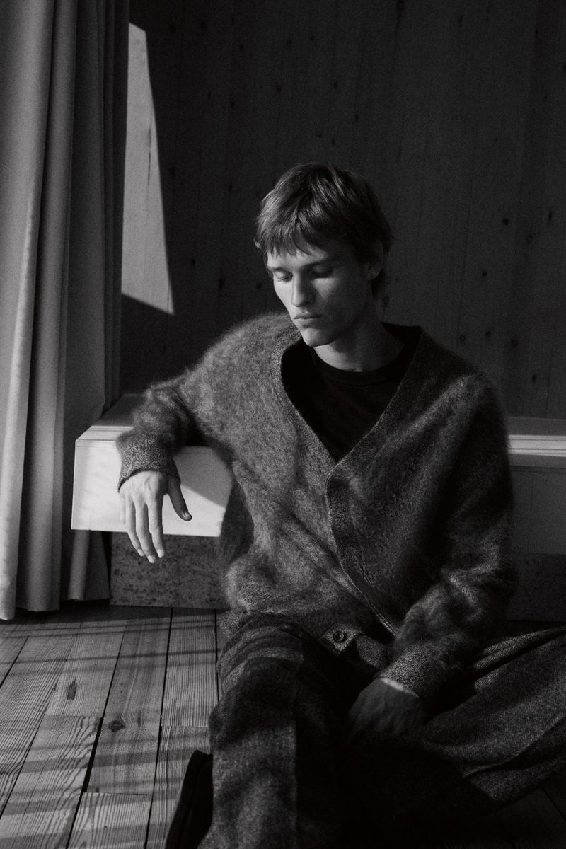 Monochrome portrait of a young man wearing a cardigan, seated on a wooden floor, next to a window with light and shadows.
