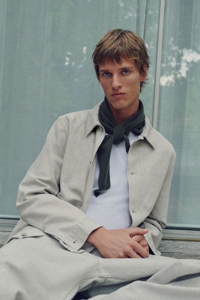 Portrait of an adult male with light brown hair wearing a light-colored jacket and a dark scarf sitting outside against a glass wall.