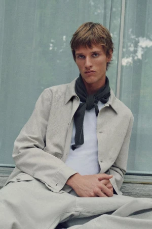 Portrait of an adult male with light brown hair wearing a light-colored jacket and a dark scarf sitting outside against a glass wall.