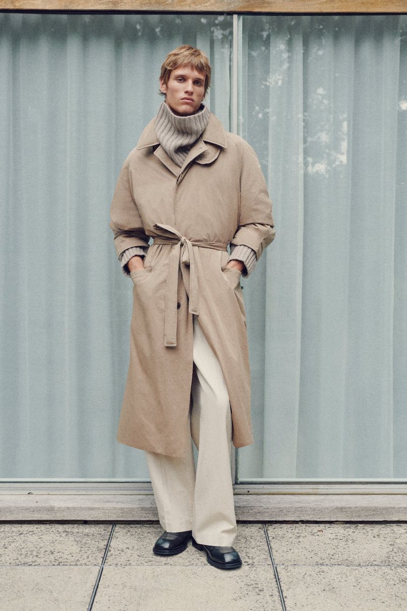 A blond male model stands in a long tan coat with a tie belt, white pants, and black shoes, against a blue paneled background.
