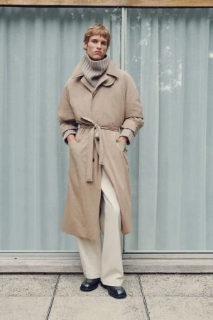 A blond male model stands in a long tan coat with a tie belt, white pants, and black shoes, against a blue paneled background.