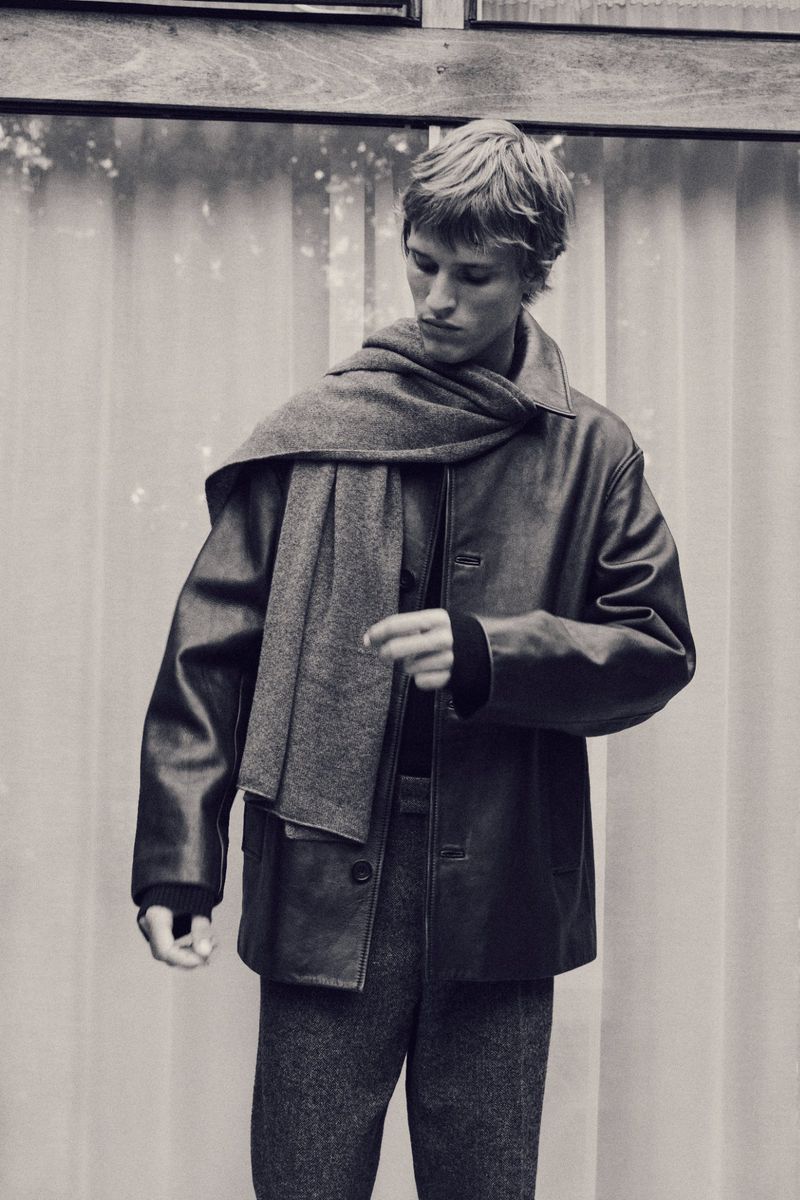 Portrait of a blonde adult male fashion model wearing a dark leather jacket and grey scarf in front of a bright window.