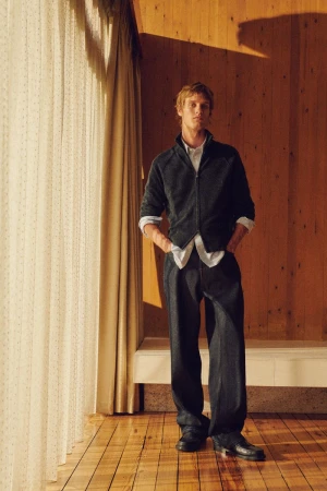 Man in a dark charcoal zip jacket and matching wide-leg trousers standing beside sheer window curtains on wooden flooring.