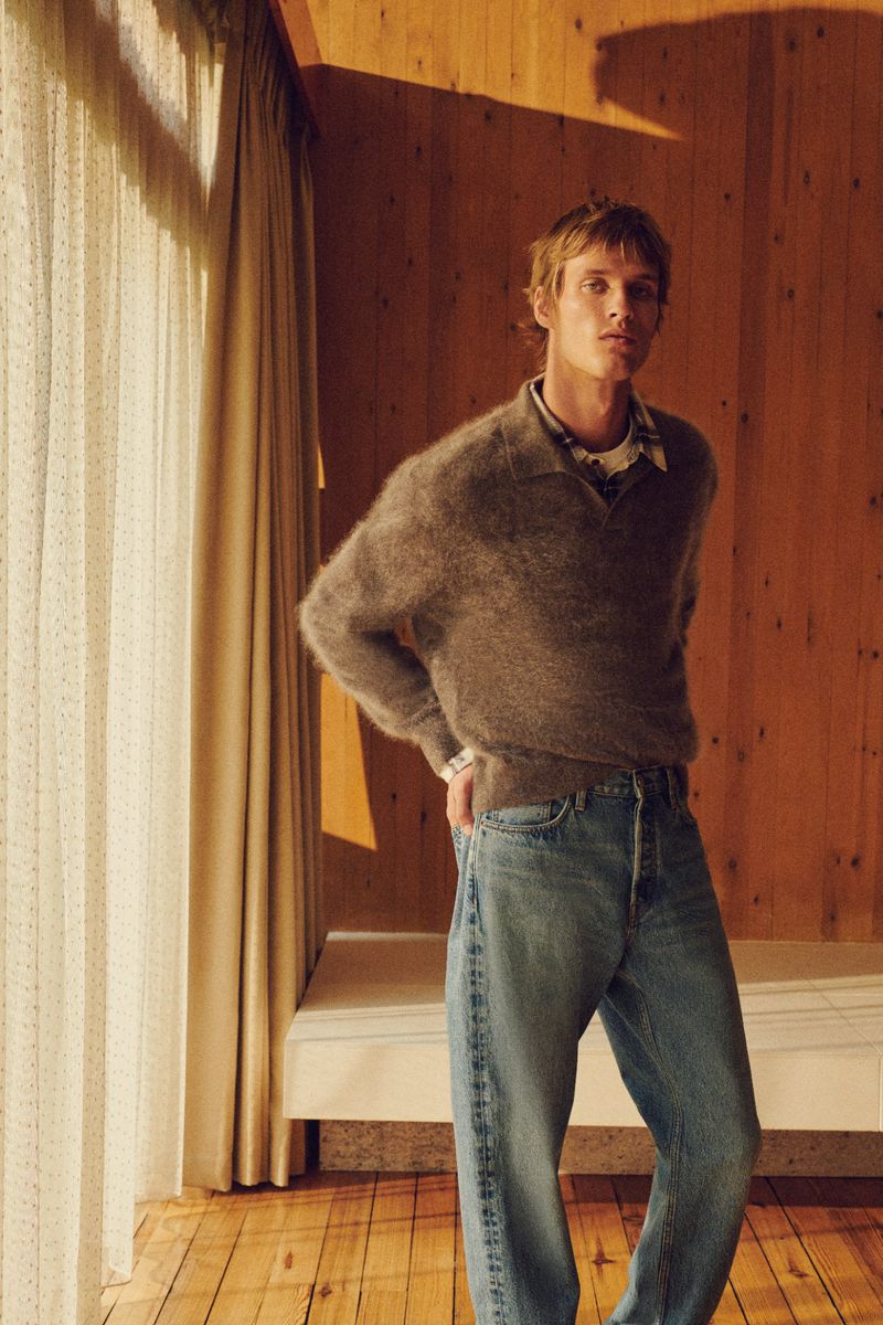 Adult male in a fuzzy brown sweater and jeans, standing indoors near a window. The background features light wood paneling.